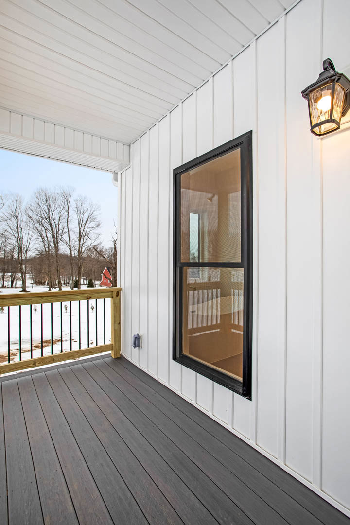 Wooden porch with railing, screen door, window, and lamp overlooking snowy landscape and trees