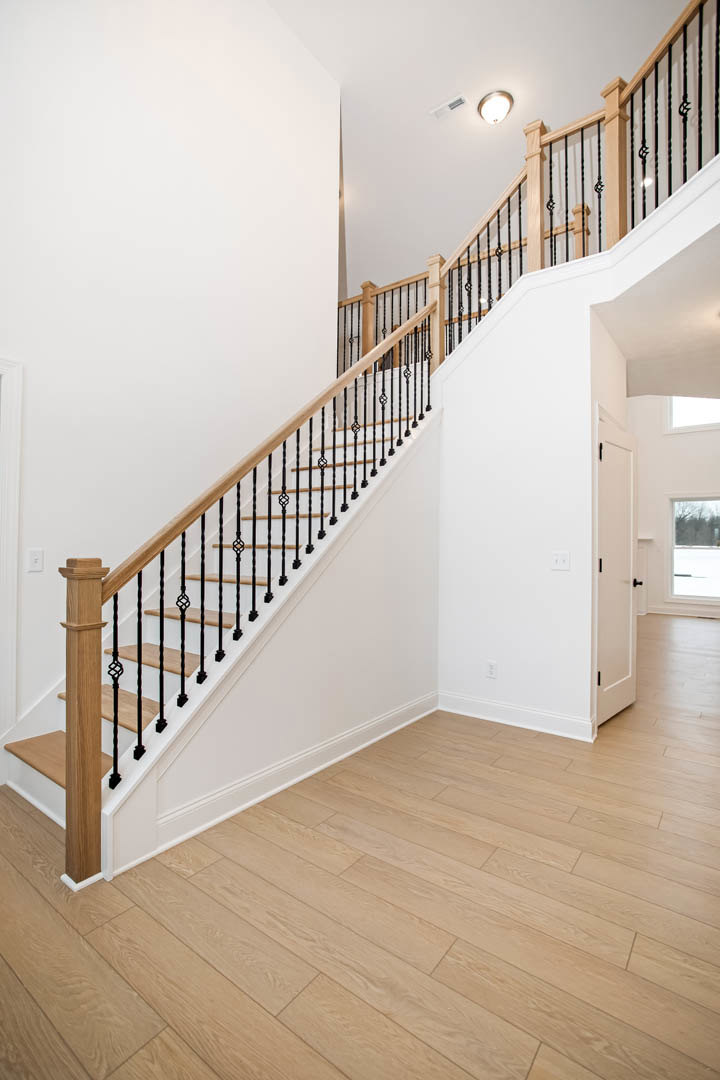 Wood staircase with white balusters and wooden handrail, set on light hardwood flooring; large window with tree views in background, ceiling light fixture above.