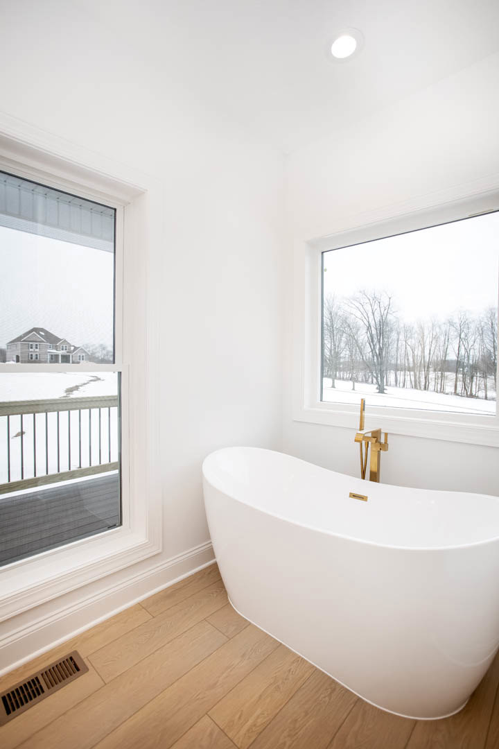Freestanding white bathtub beside large window overlooking snowy landscape, wood floor with vent, outdoor deck visible through glass railing, bird flying outside