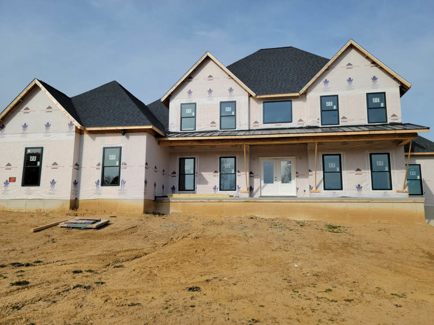 Partially built house with exposed framing, black-framed windows, construction tape along exterior walls, dirt lot in foreground, blue sky with scattered clouds