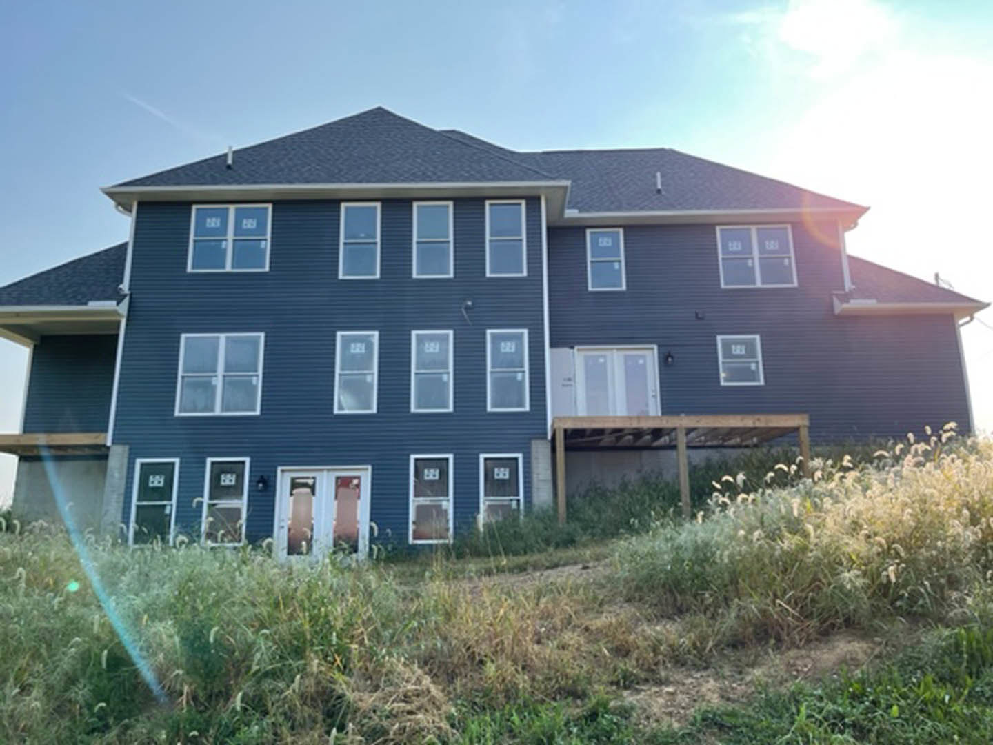 Two-story home with large windows, wooden porch, and deck, set on grassy lawn with a hill and blue sky in the background