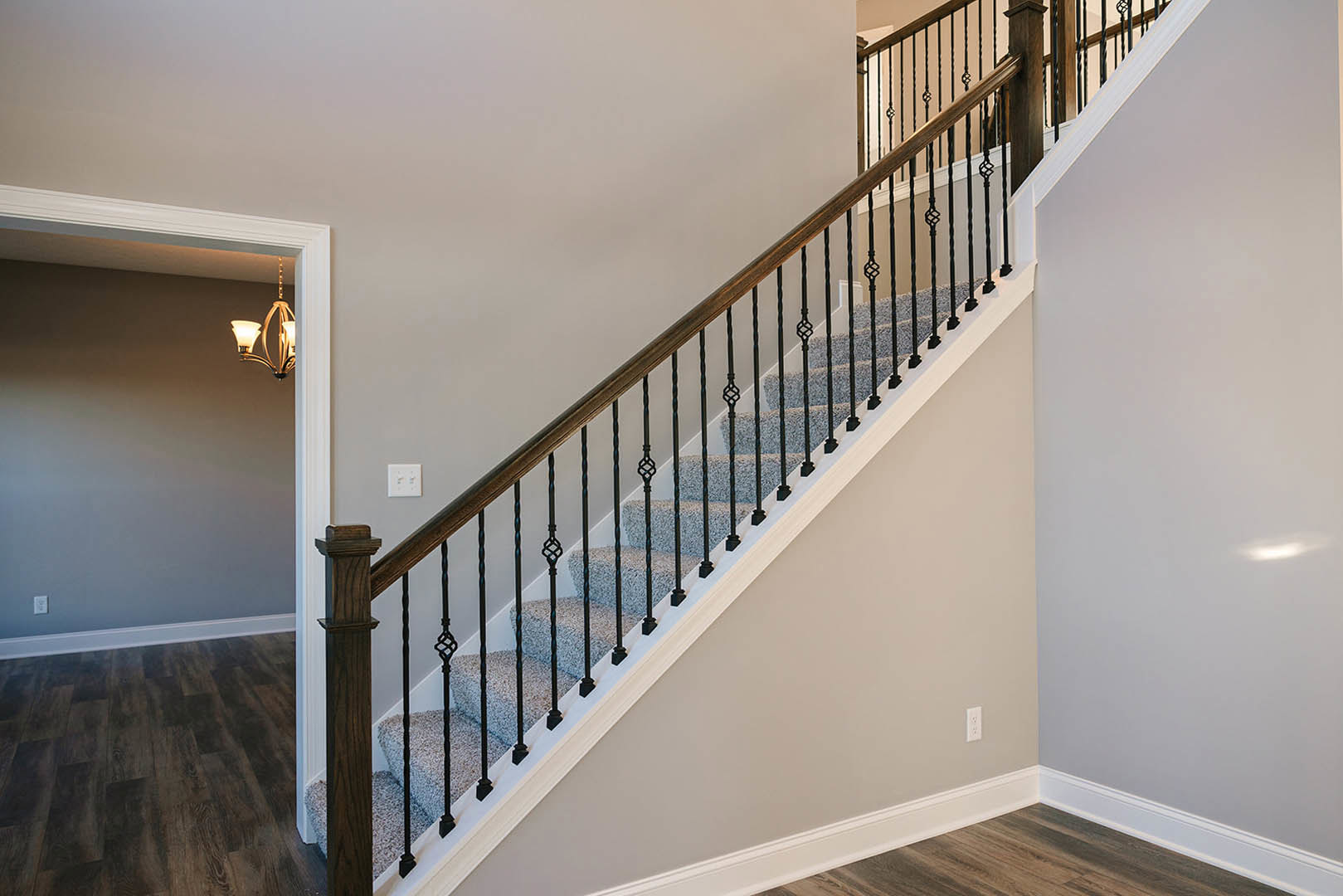 Wood staircase with black metal railing, white walls and trim, wood flooring, wall outlet with two switches, close-up of wood paneling and chandelier
