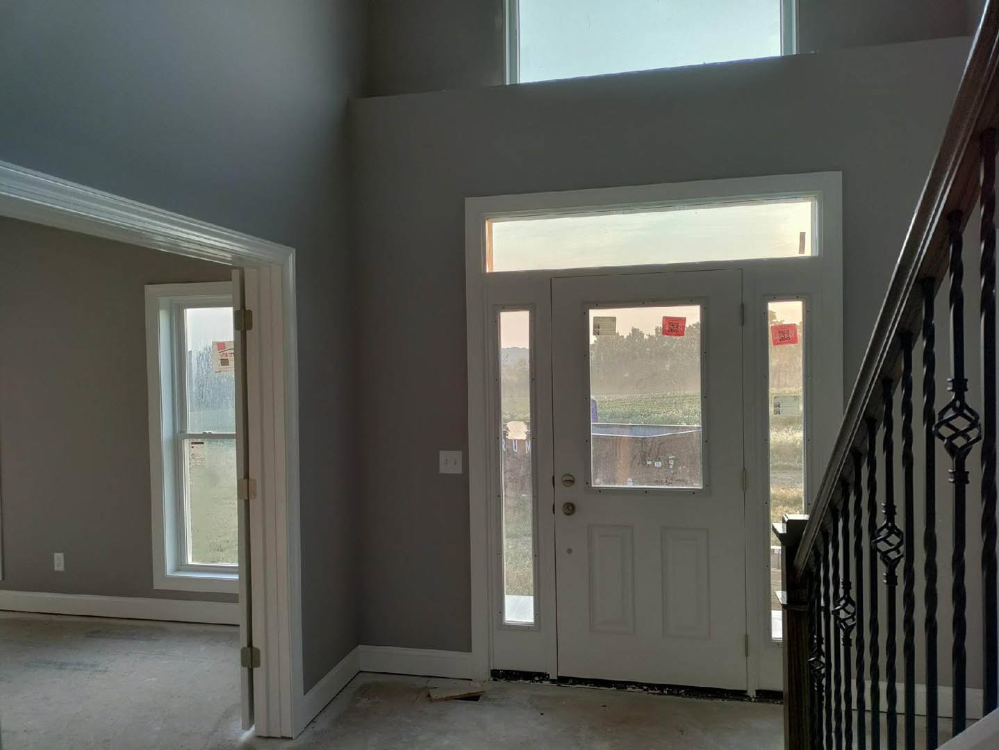 Bright room with white plaster walls, wood flooring, large windows, and a modern door; staircase with metal handrail visible, red signs posted on window.