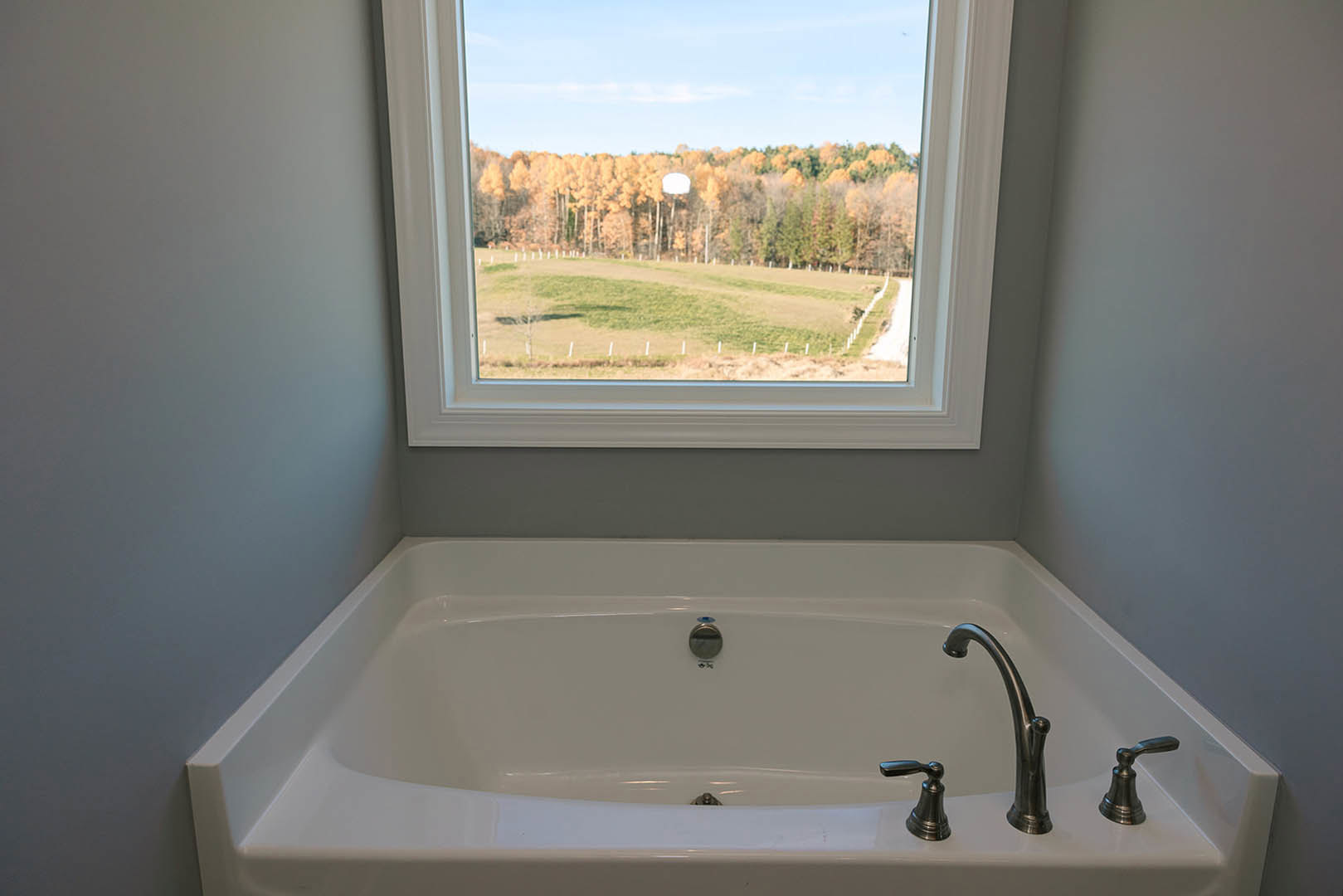White freestanding bathtub with silver faucets beneath a large window overlooking a grassy field and trees