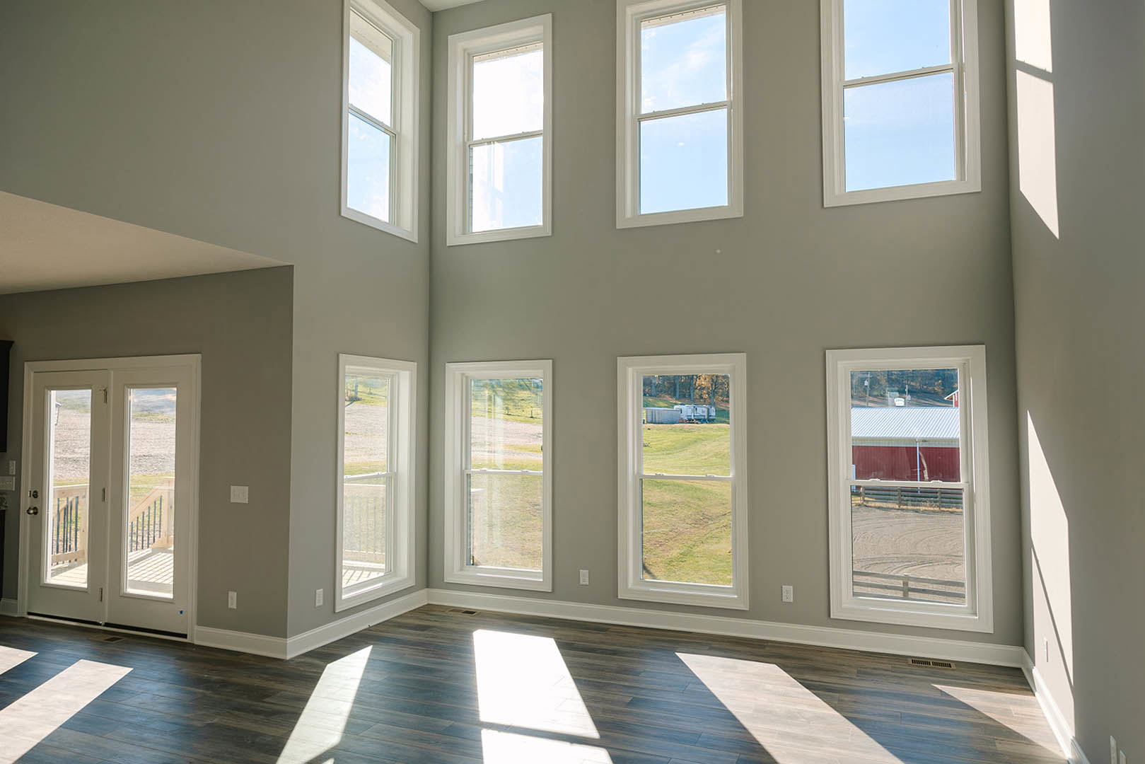 Spacious room featuring multiple large windows, sunlight illuminating light wood flooring, glass-paneled door, views of grassy yard, distant trailer, fence, and barn, white walls