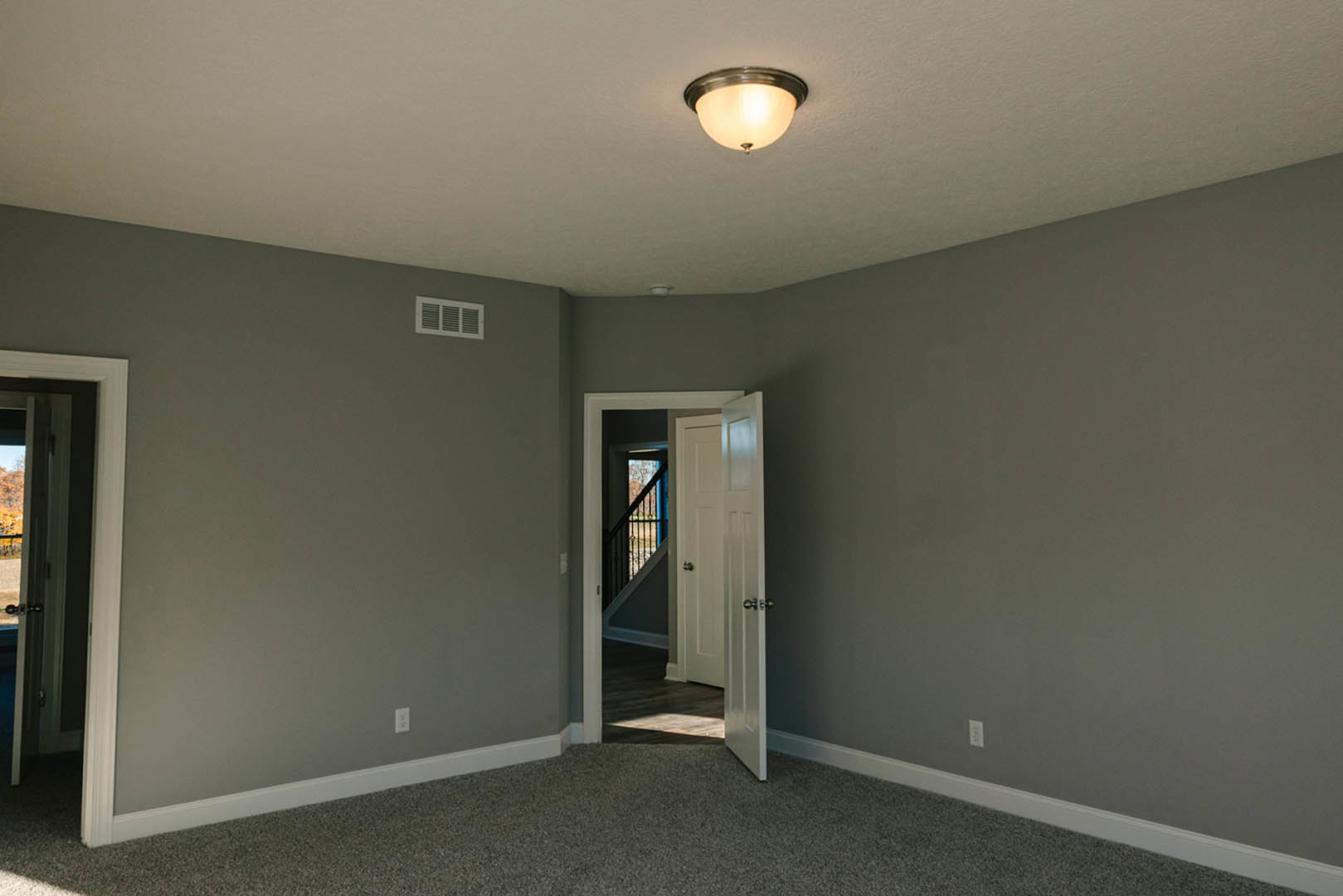 Carpeted room with white walls, ceiling light fixture, open door revealing staircase, wall vent, and plaster finishes