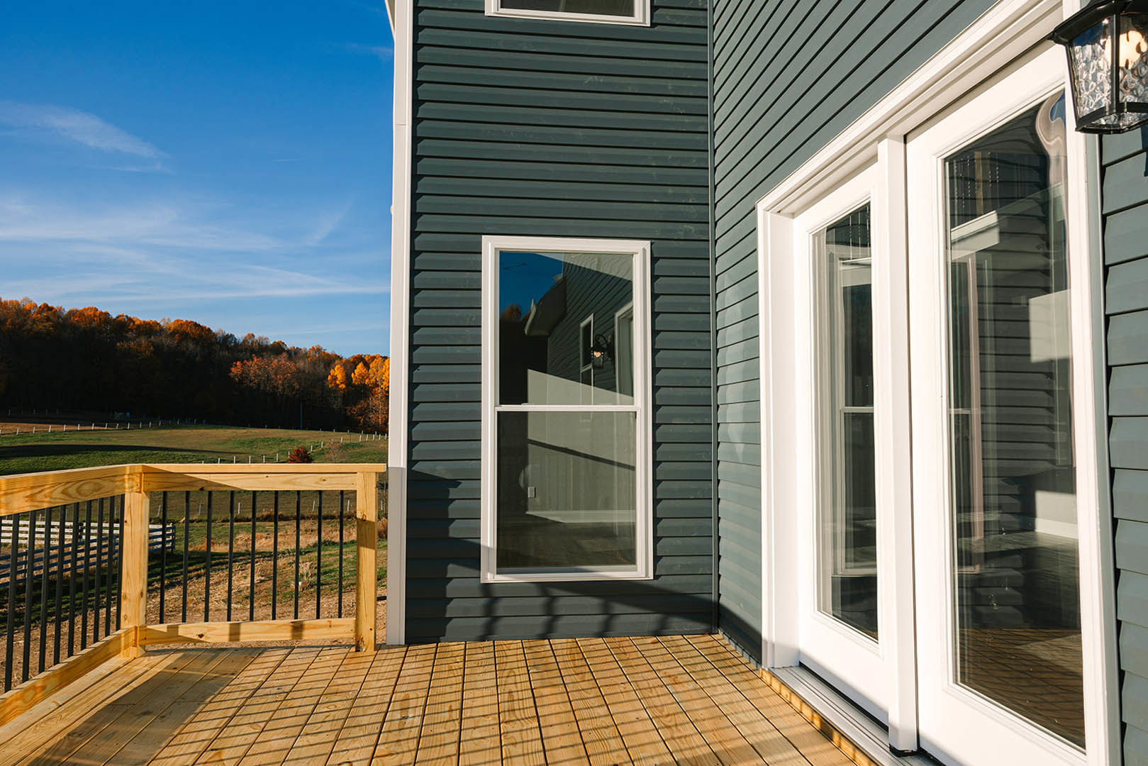 Wooden deck with black railings, white-framed window, light fixture, fenced yard, and trees with orange autumn leaves