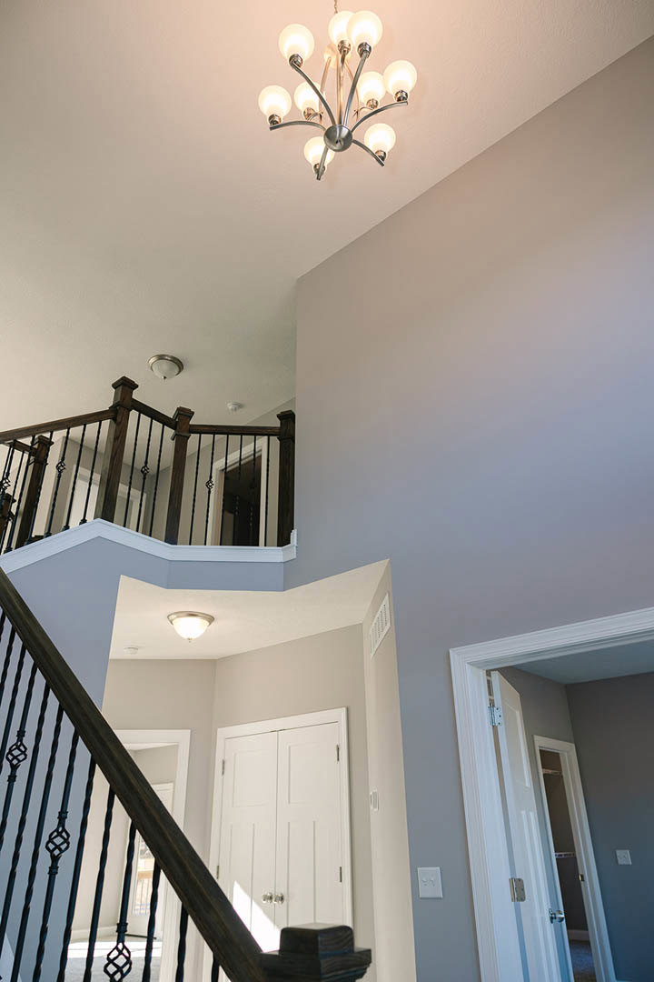 Curved wooden staircase with white balusters, dark handrail, and chandelier overhead; white paneled door with silver handles; light gray walls with crown molding