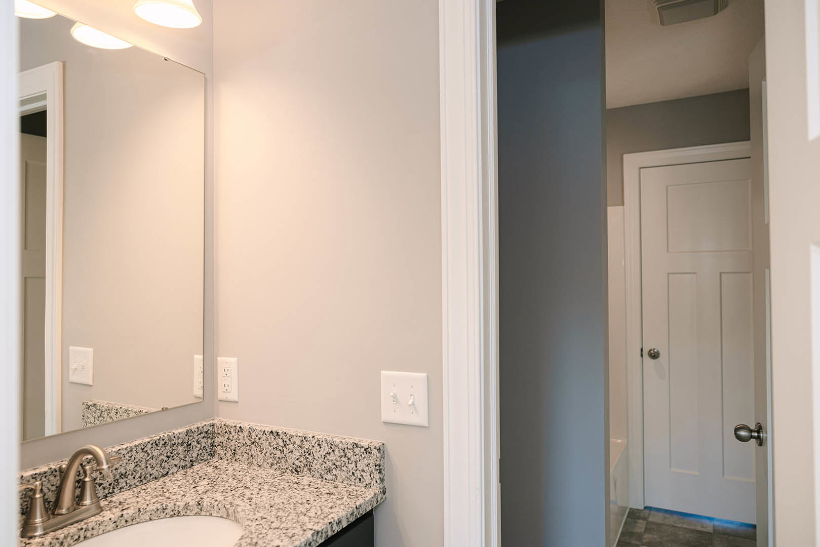 White bathroom with rectangular mirror above undermount sink, chrome faucet, light gray stone countertop, white tile backsplash, and double light switch on painted wall