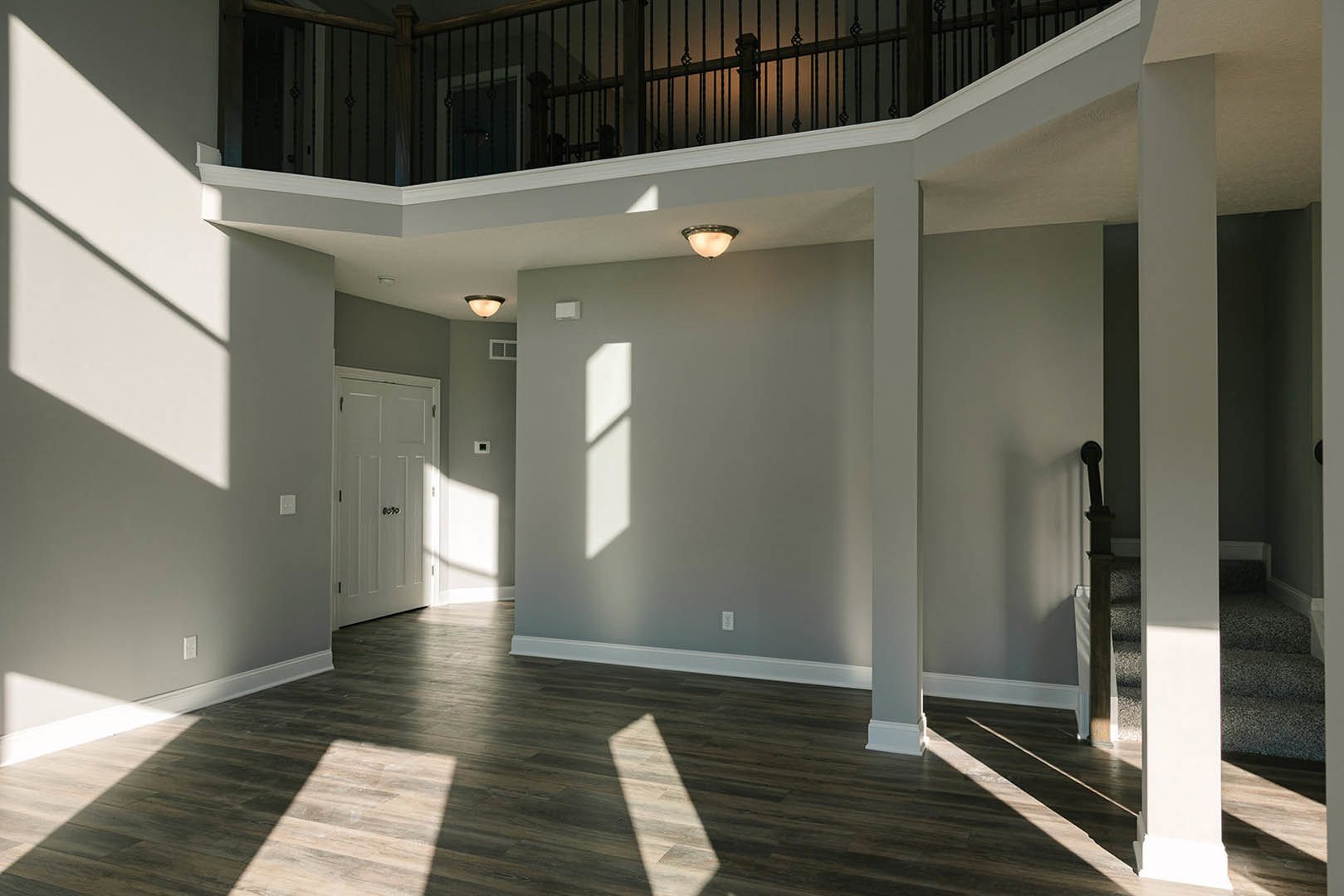 Open-concept room featuring a wooden staircase with black metal railing, white door with black handles, warm light illuminating hardwood floors, and a black pillar near the