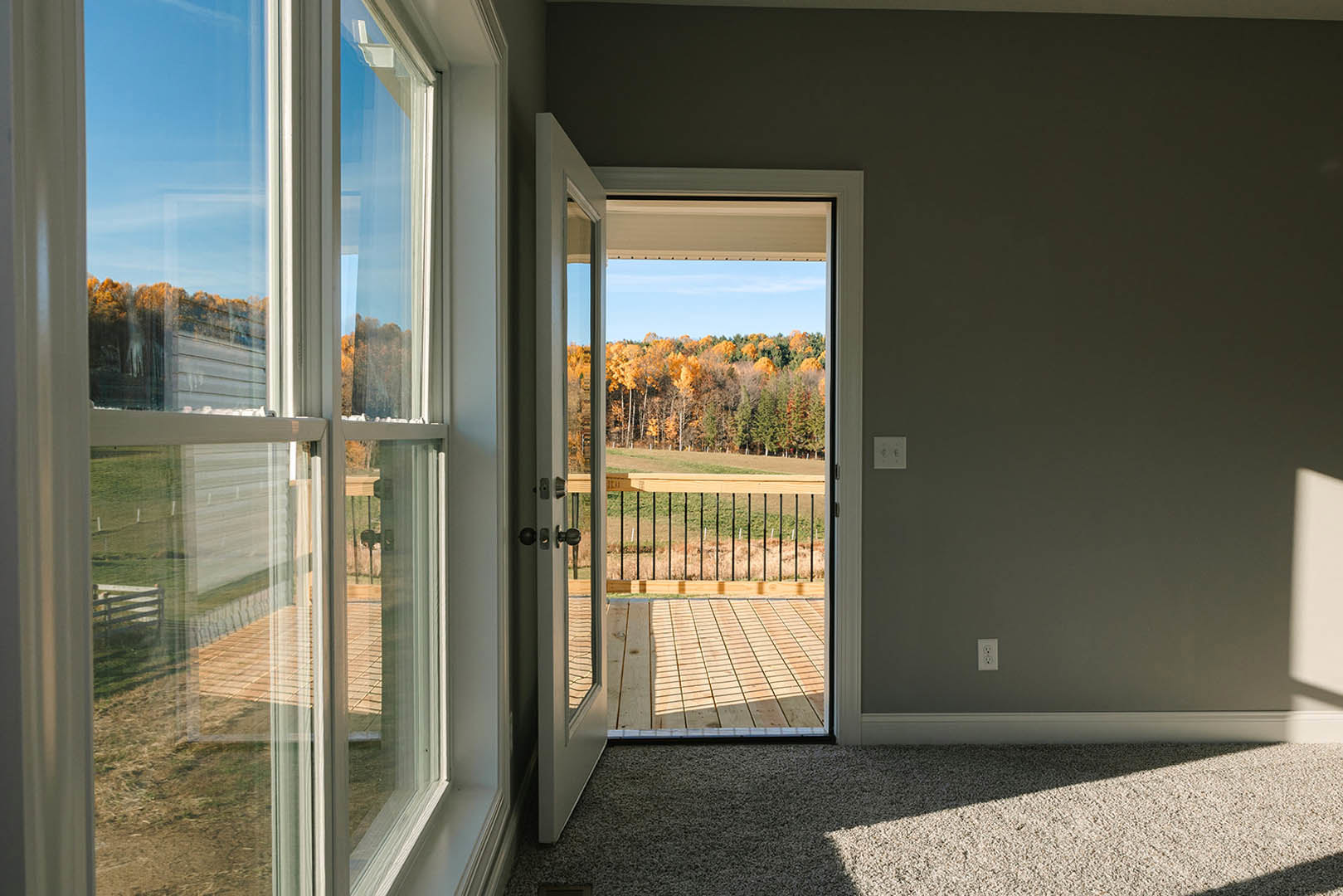 Carpeted room with open door leading to wooden deck, forest view through window, wood railing and fence visible outside