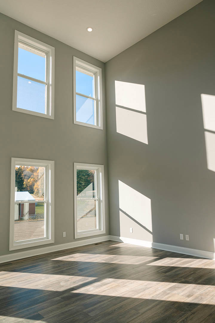 Sunlit room with wide windows, white frames, and natural wood flooring; blue sky visible outside, window shadows cast across the floor.