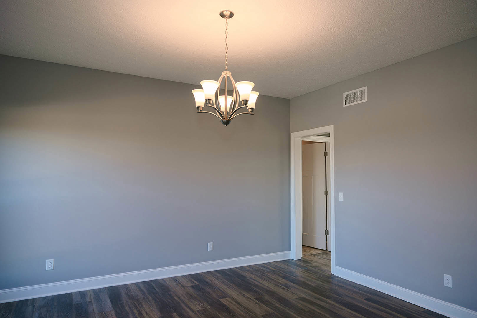 Chandelier with glass accents hangs from white ceiling above wood floor; white baseboards line pale walls, black-handled door and wall vent visible.
