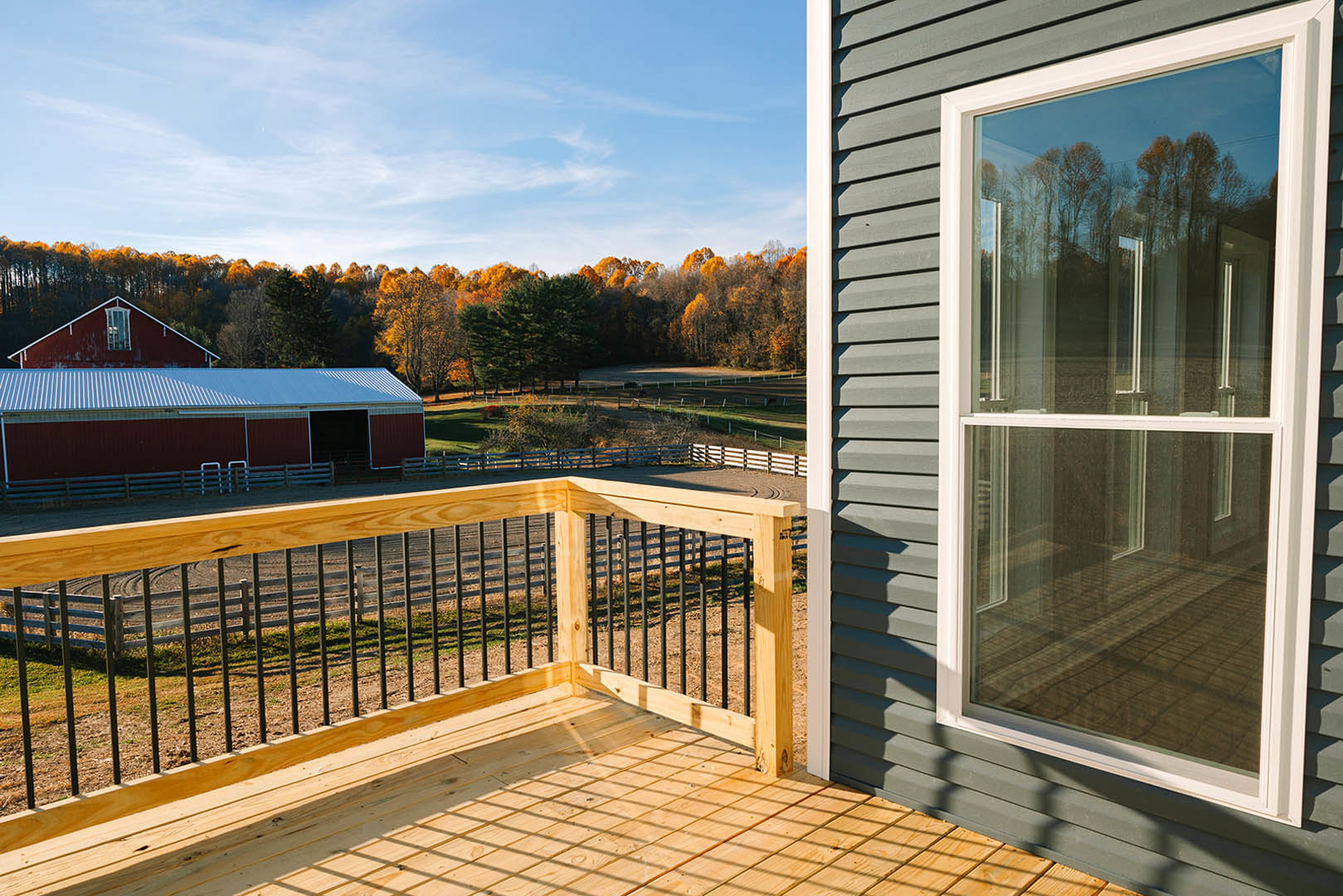 Wooden deck with metal railing overlooking a red barn with white trim, surrounded by trees and sky, window visible on adjacent house exterior