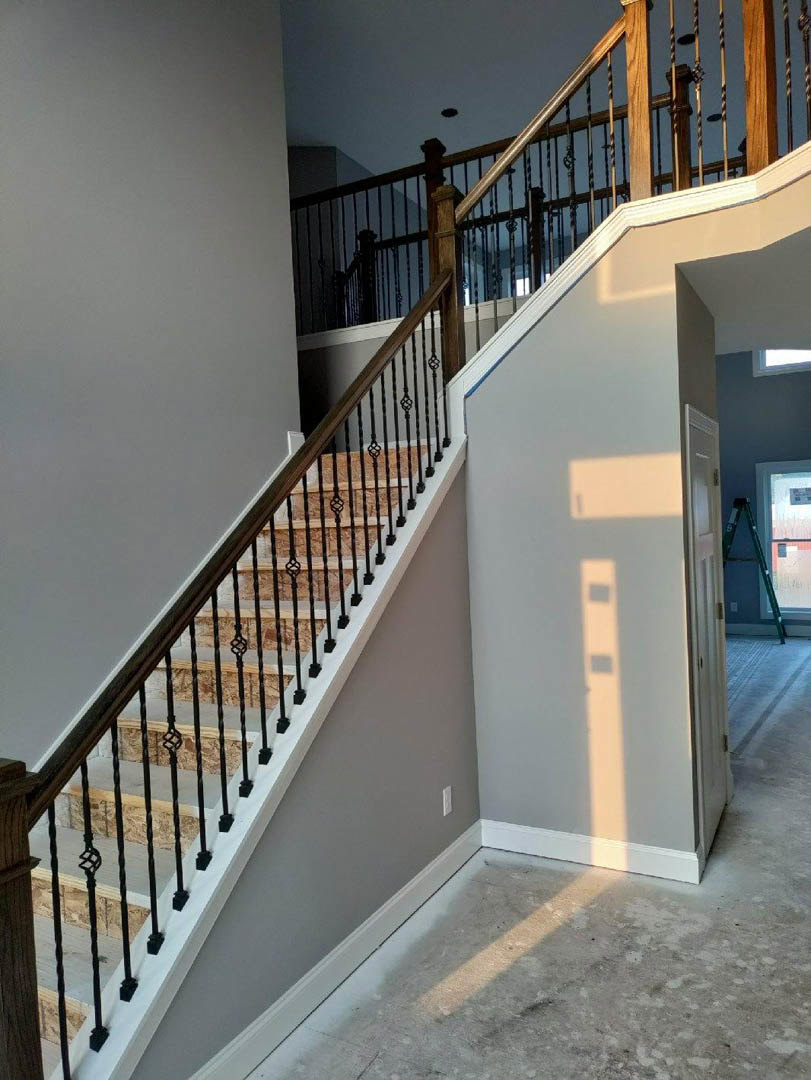 Wood staircase with black metal railings, square window with black handle, sunlight casting door shadow on white wall