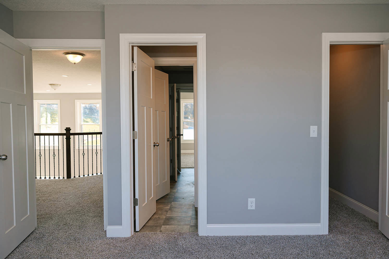 White hallway with wood flooring, metal railing, white doors, wall-mounted light fixture, and brushed metal door handle