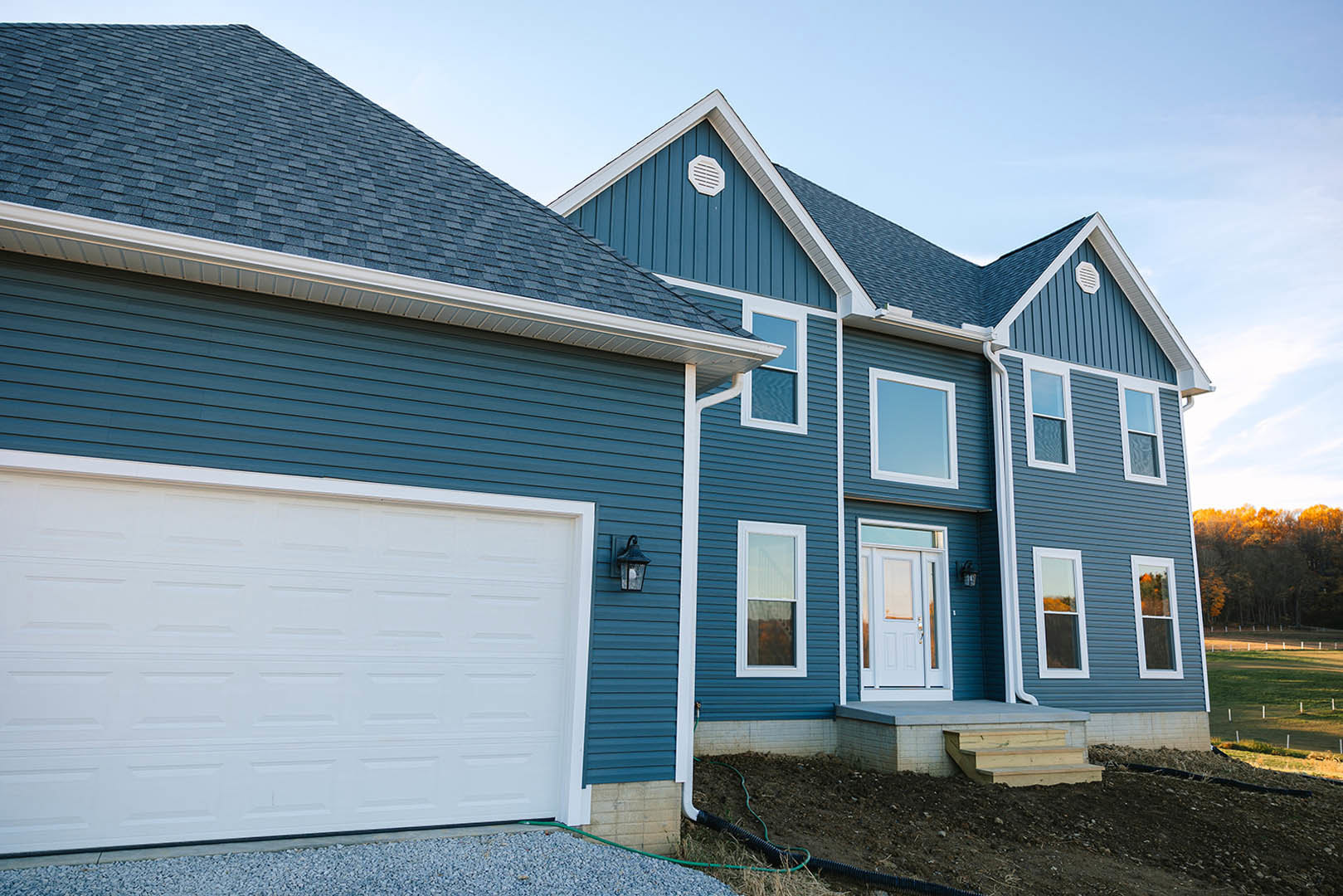 Blue siding exterior with white garage door, glass-paneled entry door, white vent, and window; porch area visible under clear sky.