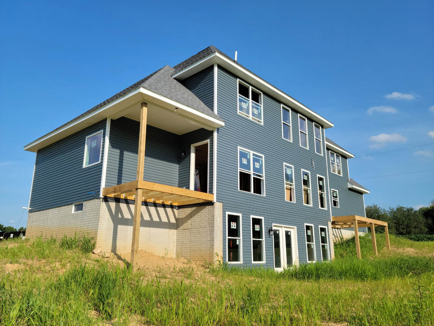 Wood-framed house under construction with exposed beams, open porch, grassy yard, blue sky, and visible windows and door.