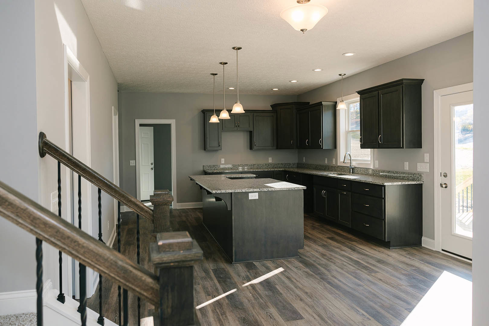 Kitchen with dark wood cabinets, granite island countertop, stainless steel sink, staircase with wooden steps, pendant light fixture, hardwood flooring, white walls, and modern