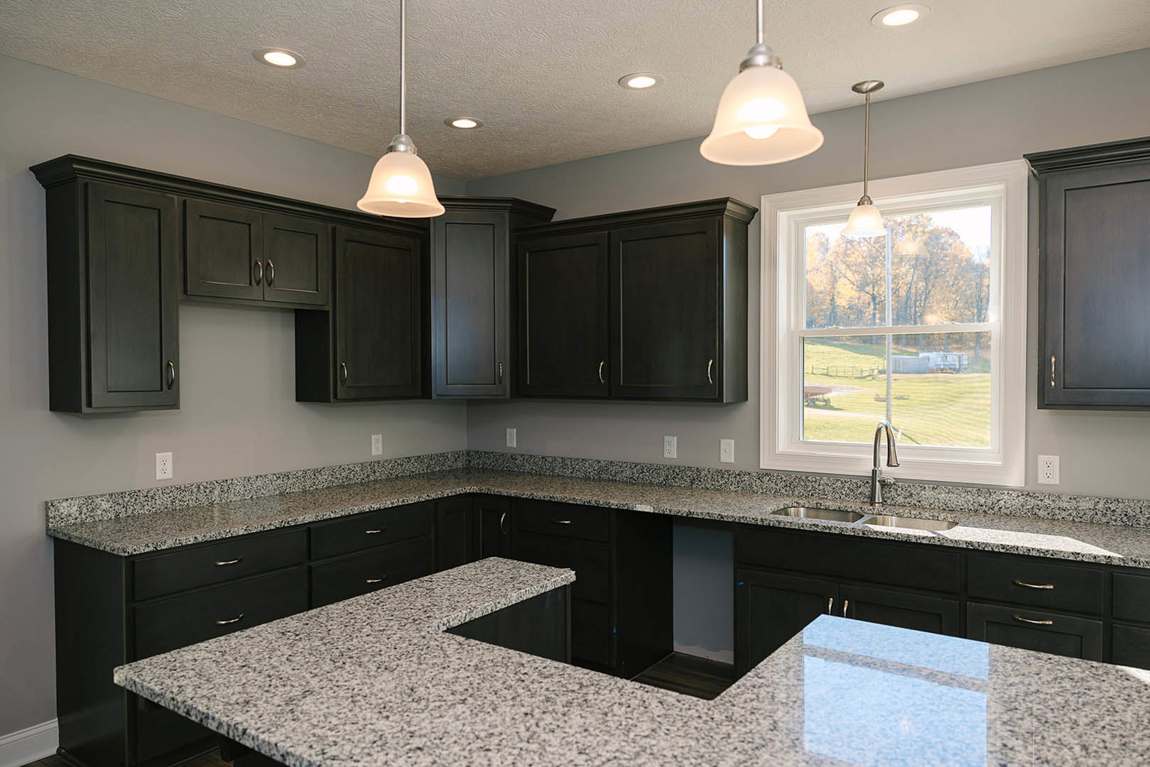Granite countertops, tile backsplash, stainless steel sink, black cabinetry, window overlooking grassy field, recessed lighting, grey painted walls