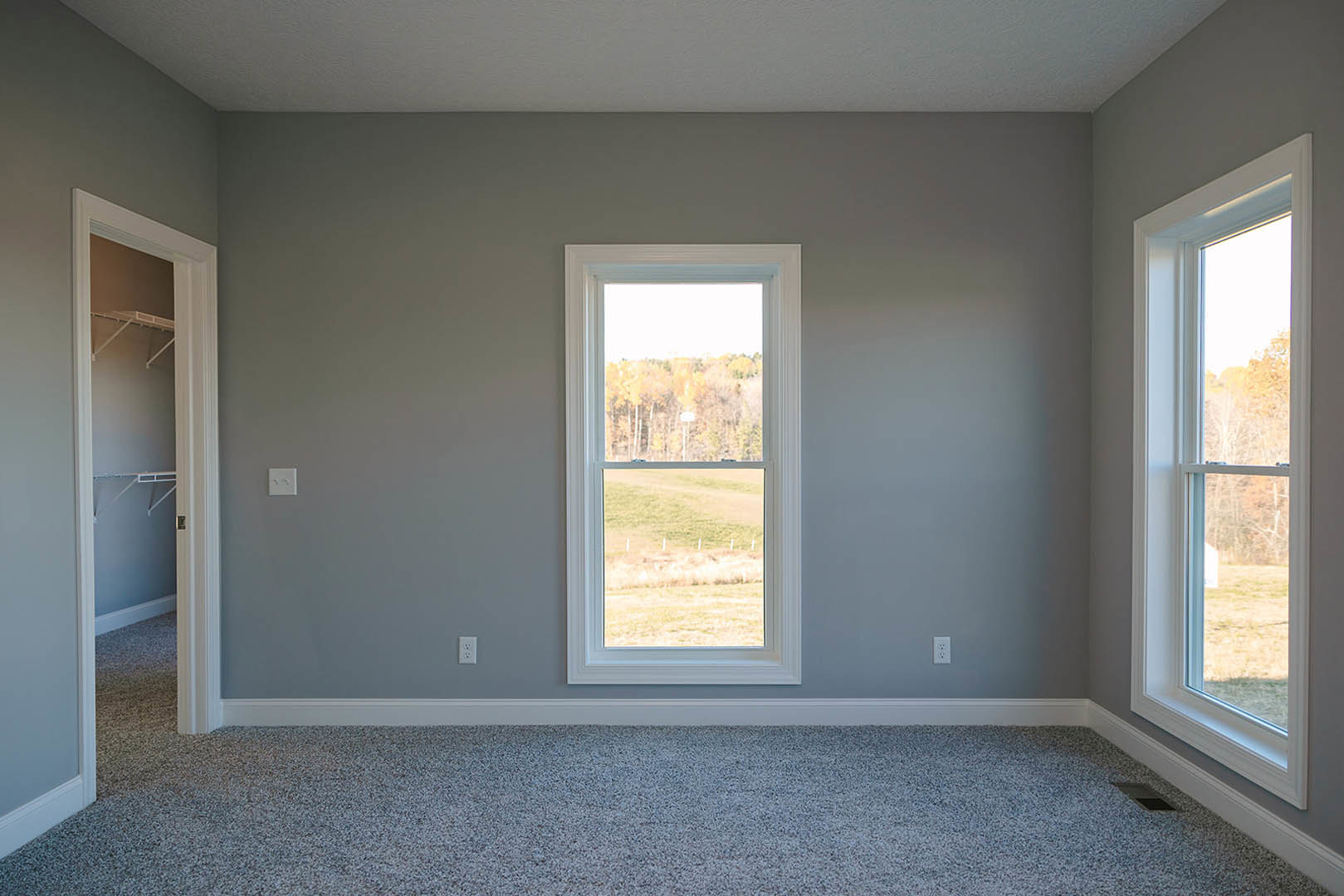 Large window with white trim overlooking green trees, beige carpet flooring, white walls, and minimal decor in a bright residential room