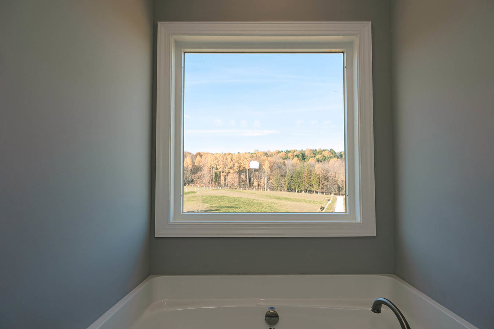 Large bathroom window with tree views above freestanding white tub, chrome faucet, and light-colored wall finishes