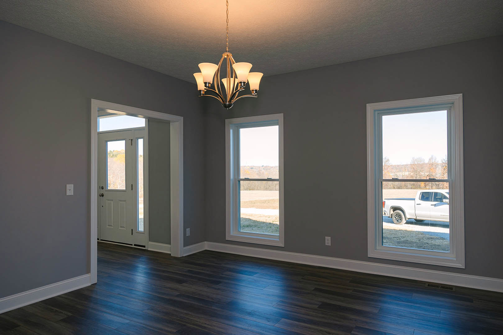 Chandelier with five lights hanging from white ceiling, dark wood floor, large windows showing parked white truck and open field outside