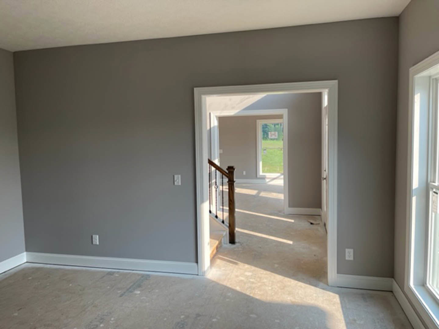 Hallway with light-colored wood flooring, white plaster walls, staircase with wooden steps and metal railing, white door, large window overlooking grassy field, wind chime hanging
