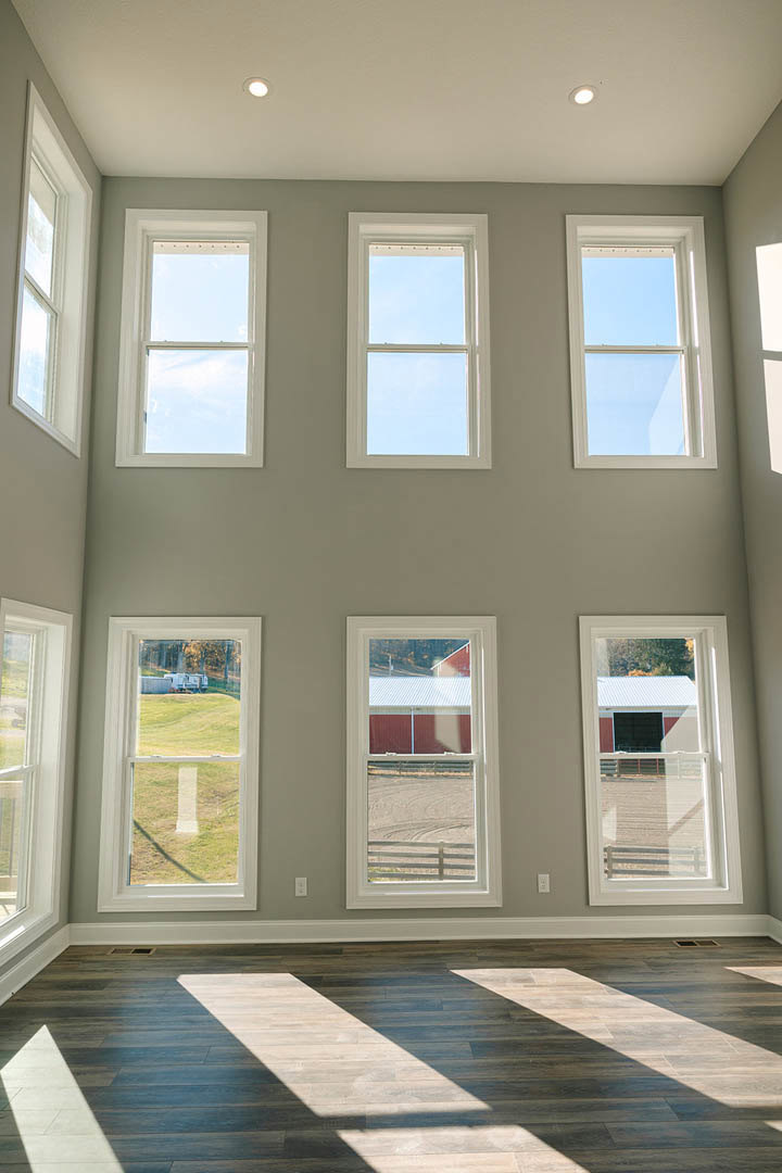 Spacious room with multiple large windows, wooden floor with sunlight and shadows, views of a truck, barn, red building, and blue sky through windows