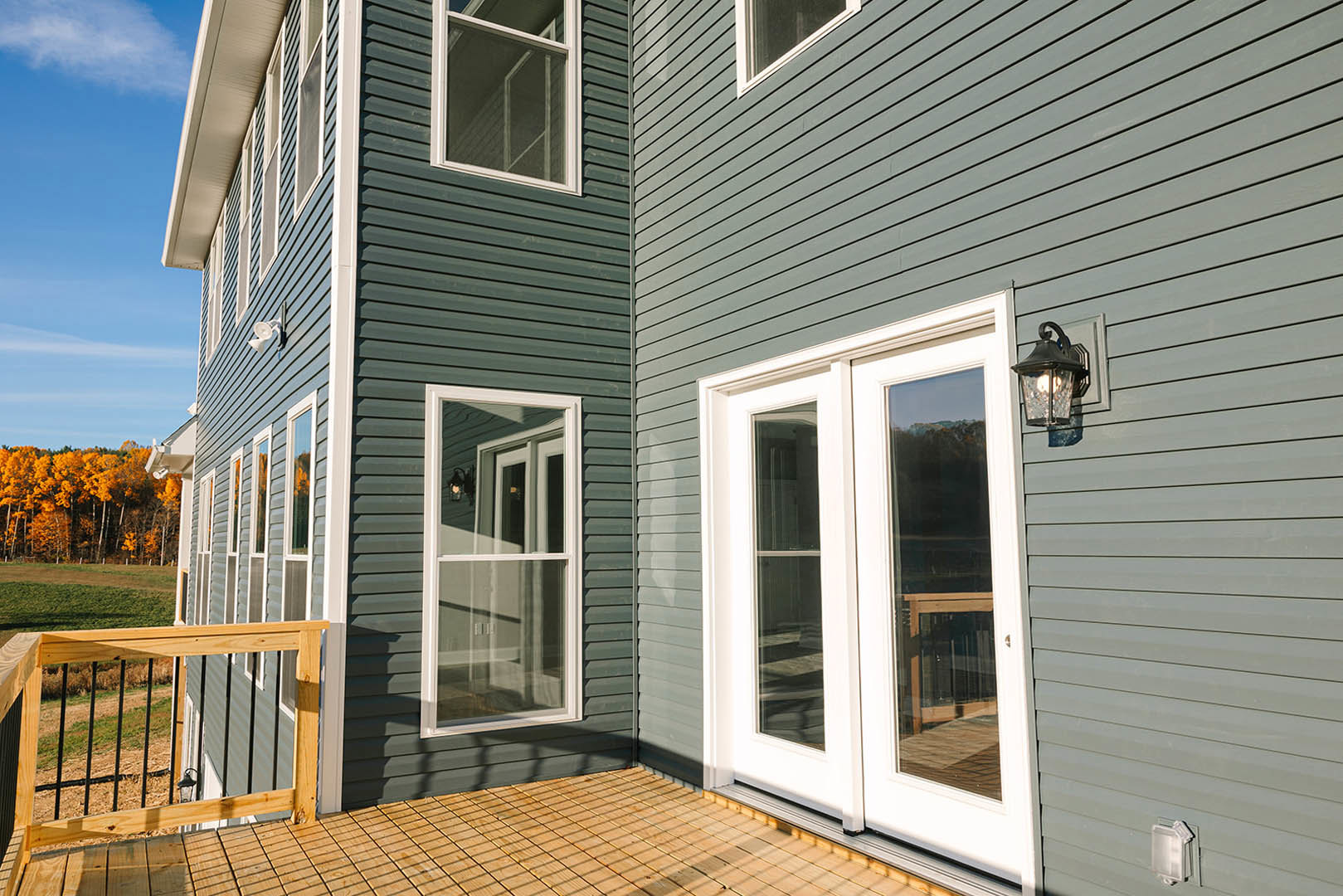 Two-story house with gray siding, wooden porch, white door, large windows, autumn trees in background, wall-mounted light fixture, white mailbox on exterior wall
