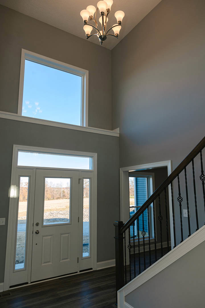 Wood staircase with white risers and railing leads to a glass-paneled white door, chandelier with multiple lights hangs from ceiling, large window reveals blue sky and field