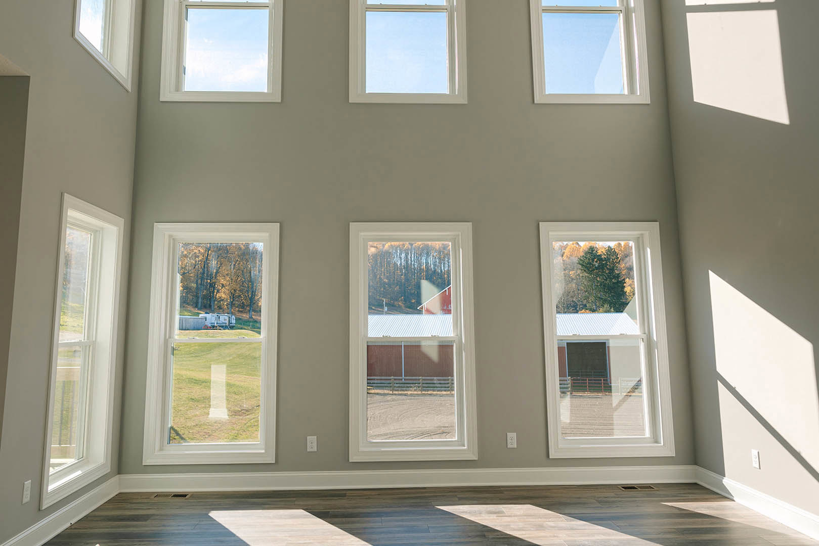 Spacious room featuring multiple large windows, wooden flooring illuminated by natural light, views of a barn, red barn, trailer, and surrounding trees visible through the windows
