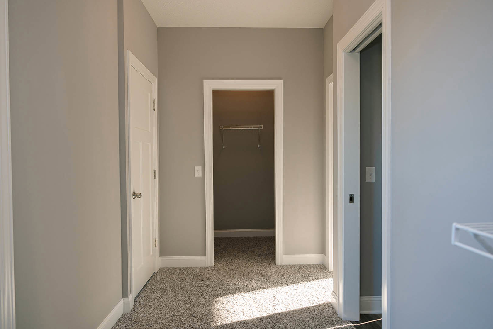 Hallway with light-colored flooring, white closet door featuring a long handle, adjacent doors, white shelving inside closet, and a light switch on the wall.