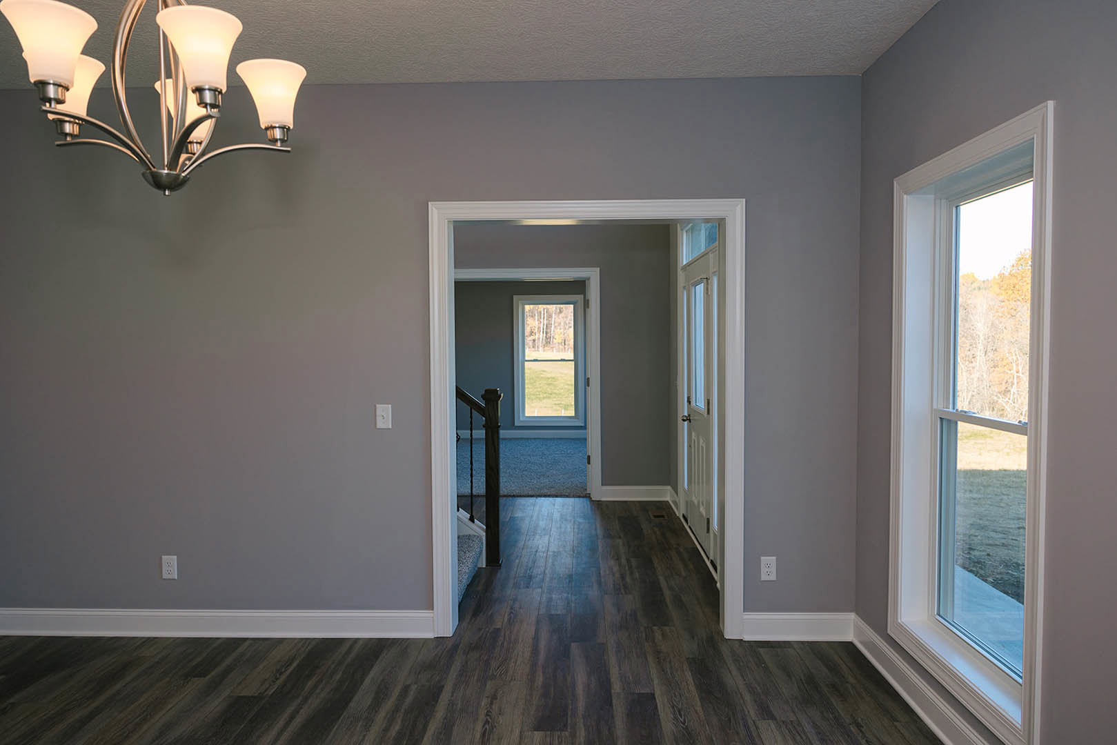 Hallway with warm wood flooring, decorative chandelier, white walls with crown molding, staircase, and large window overlooking grassy field