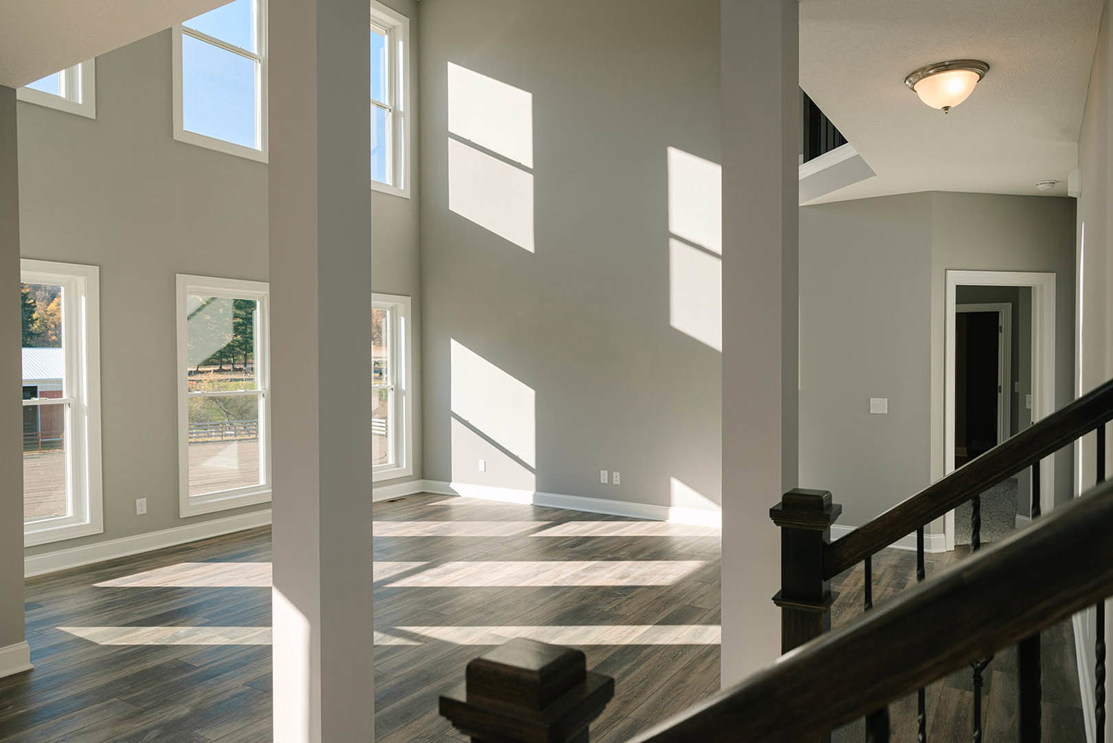 Open-plan room featuring a wooden staircase with metal balusters, large windows overlooking farmland, white walls, hardwood flooring, and a modern ceiling light fixture.