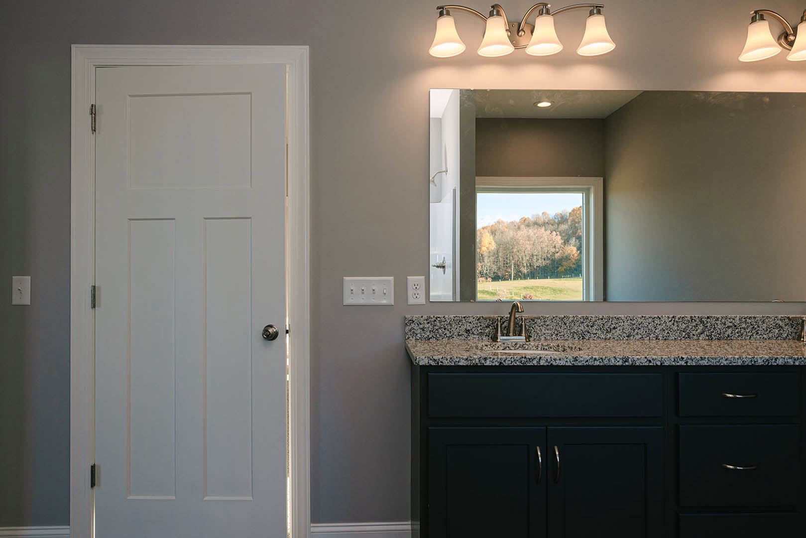Bathroom with marble countertop, rectangular mirror above sink, white door with silver knob, window showing trees outside, dual lamp light fixture, close-up of power outlet