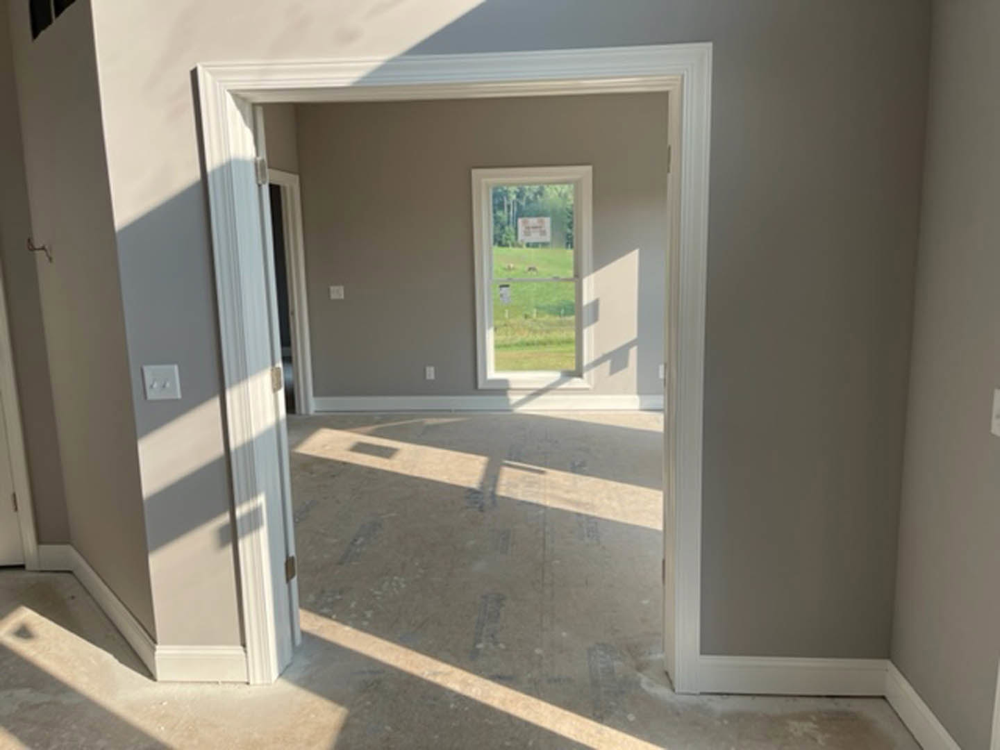 Open white door leads to a room with a large window overlooking green grass, light switch on pale wall, and floor lamp near window.