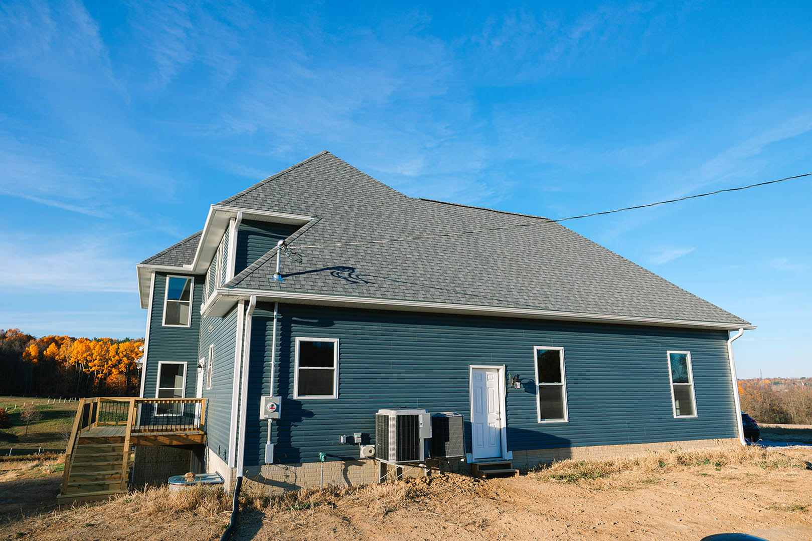 Blue siding house with white-framed window and door, wooden deck with railing, dirt path leading to entrance, satellite dish mounted on roof, surrounded by grass and trees under