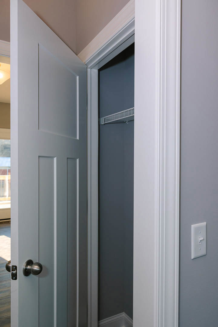 Open white closet door with silver knob, revealing adjacent room; white light switch on wall, white railing against grey wall, close-up details of door and grey wall surfaces.