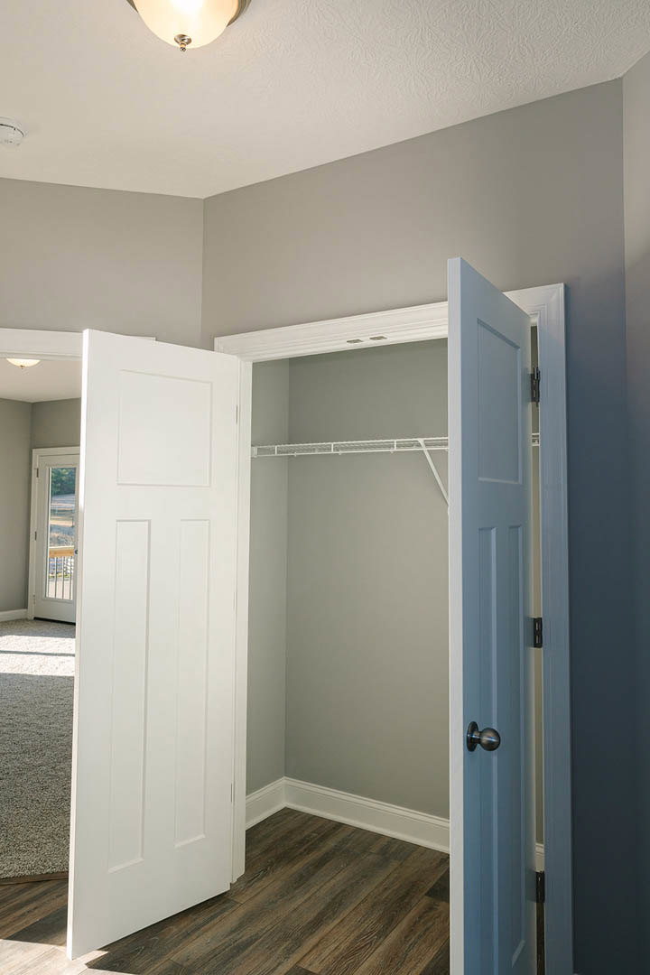 Open white closet door revealing wooden floor, white walls, ceiling light fixture, and window overlooking grassy field