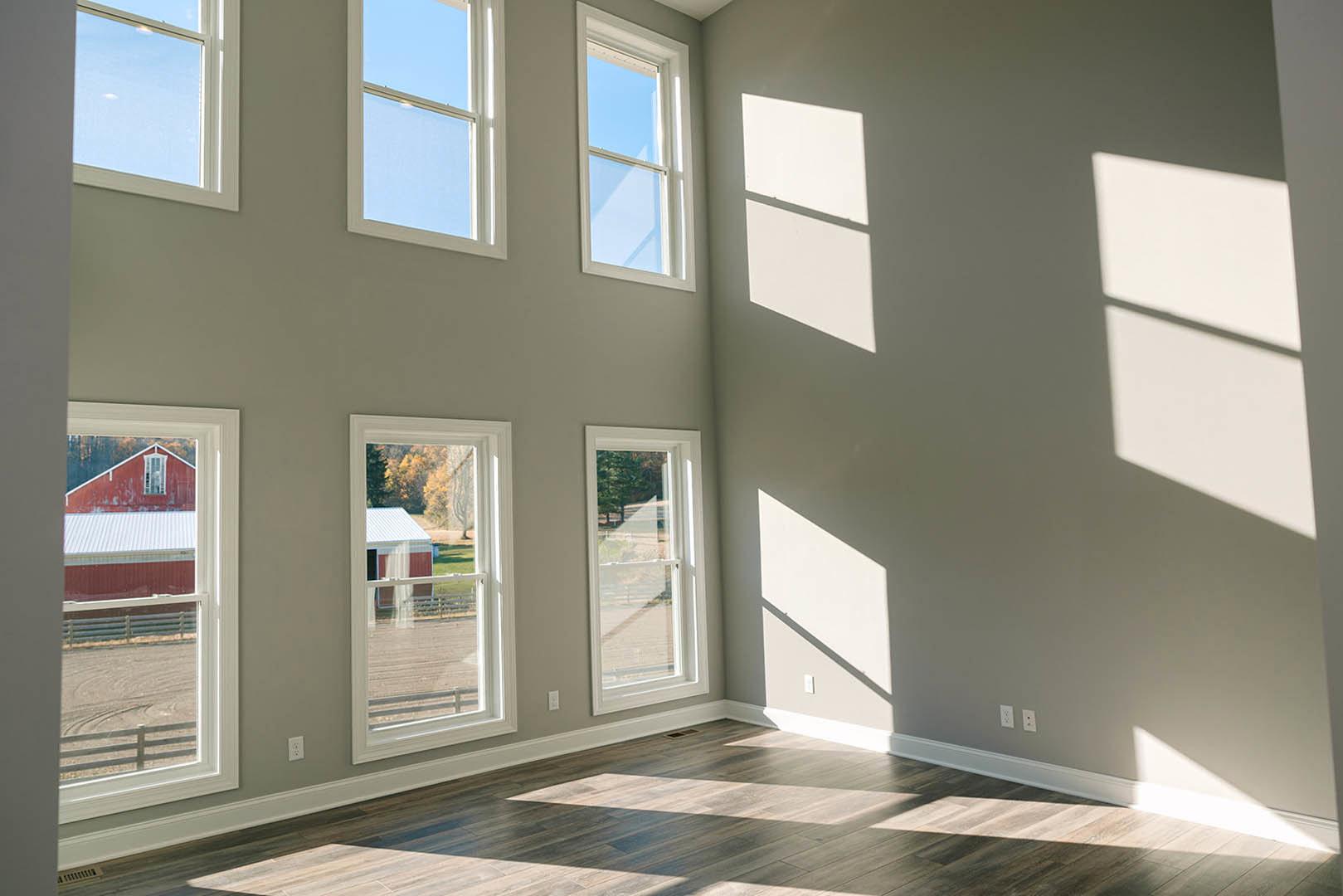 Spacious room with multiple large windows, sunlight streaming onto light wooden floors, white walls with window shadows, tree and farm visible through glass panes
