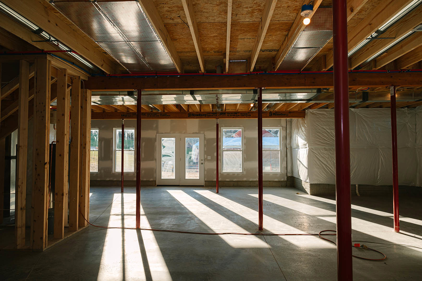 Room with red metal pole and red hose, double glass-paneled doors, white sheet draped across ceiling, exposed wooden ceiling beams, windows, and light-colored flooring.