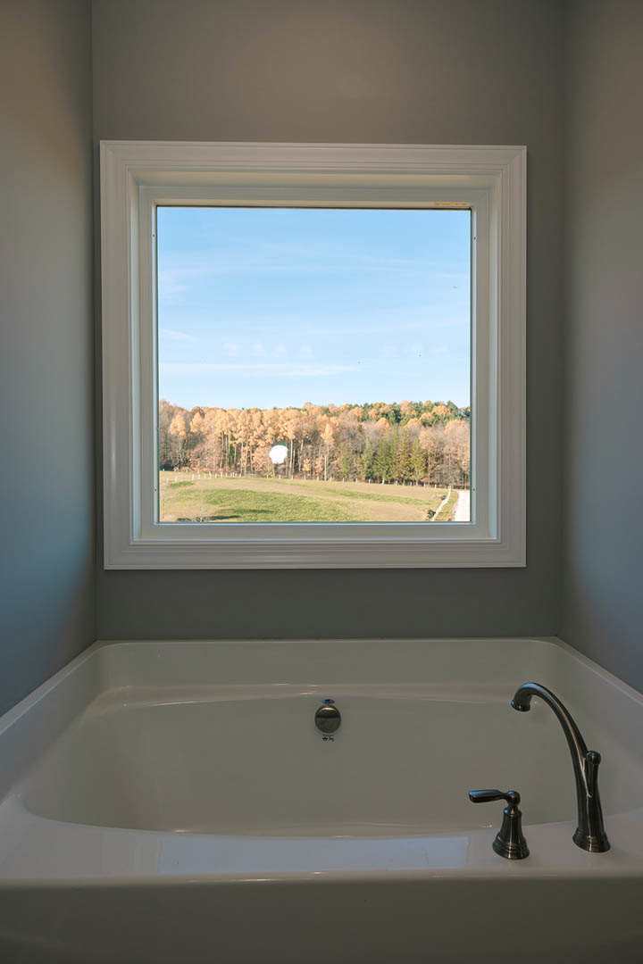 Freestanding bathtub beside large window overlooking green grass and leafy trees, chrome faucet visible