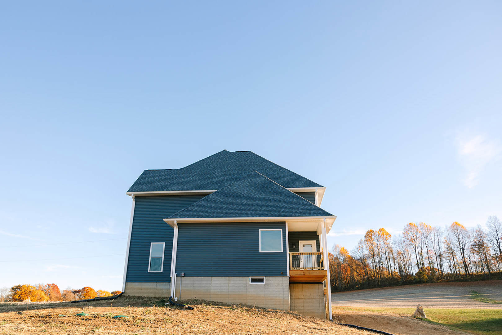 Two-story cottage with white-framed windows, covered porch, and balcony, surrounded by yellow-leaved trees, set beside a dirt road and grassy field with scattered rocks.