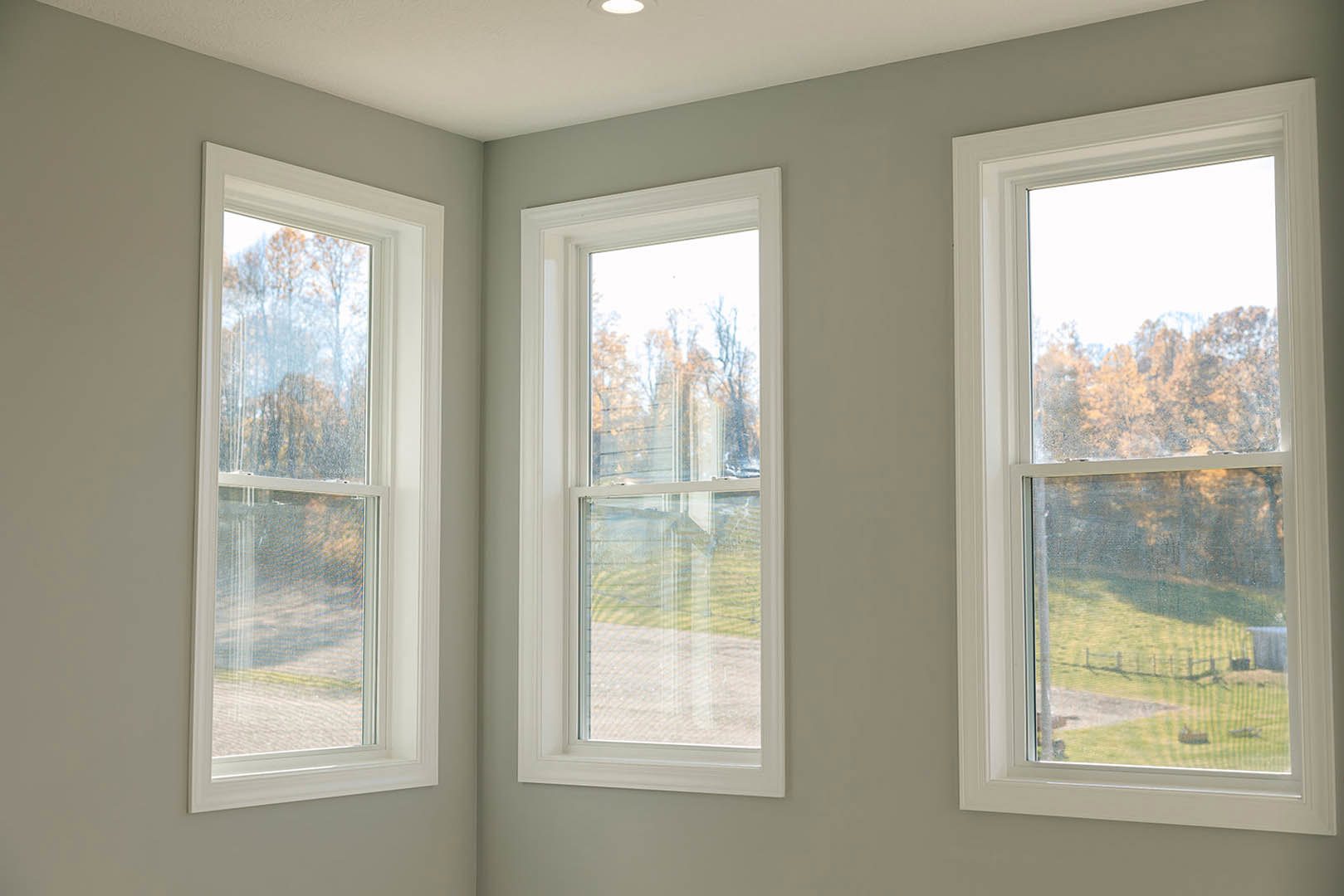 Bright room featuring three large windows with white frames, views of leafy trees outside, white ceiling with recessed light fixture, neutral walls, and minimal window blinds.
