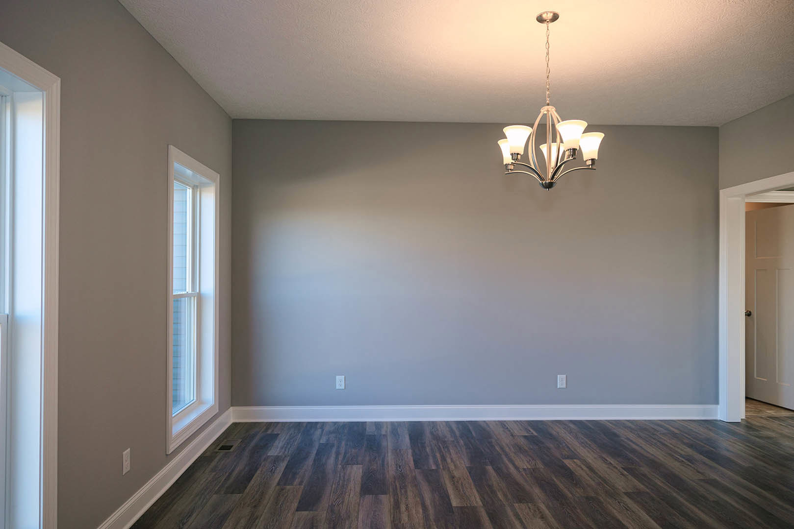 Chandelier hanging from white ceiling, wood flooring with white baseboards, large window with white trim, white plaster walls, paneled door