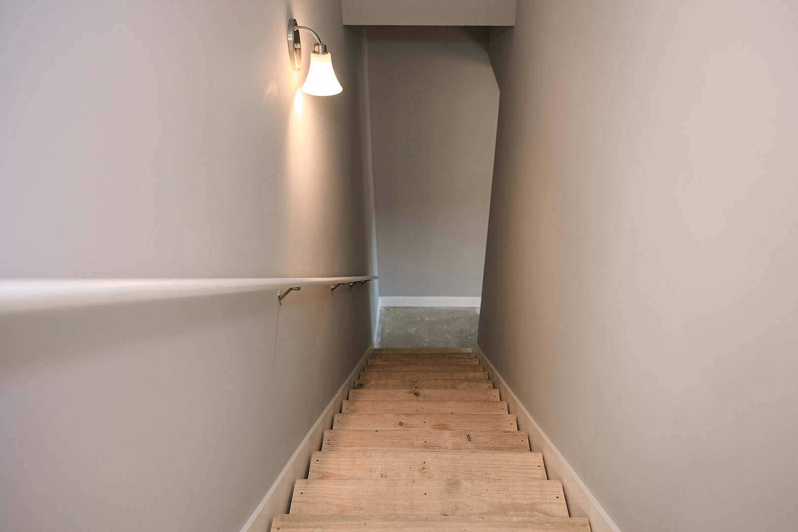 Wooden staircase with natural finish ascending to a hallway, white plaster walls with subtle shadows, close-up of a modern lamp, grey wall with white speckled surface, light