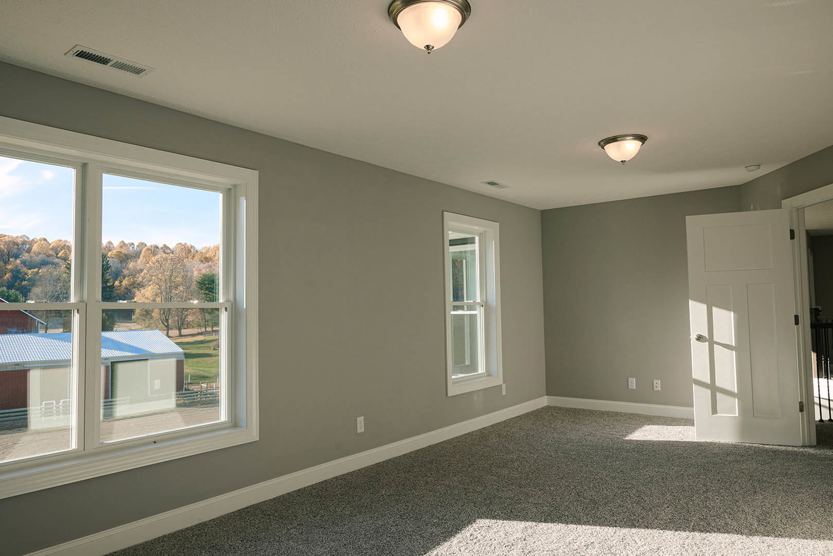 Carpeted room with multiple white-framed windows, white door with silver handle, and view of farm and trees outside