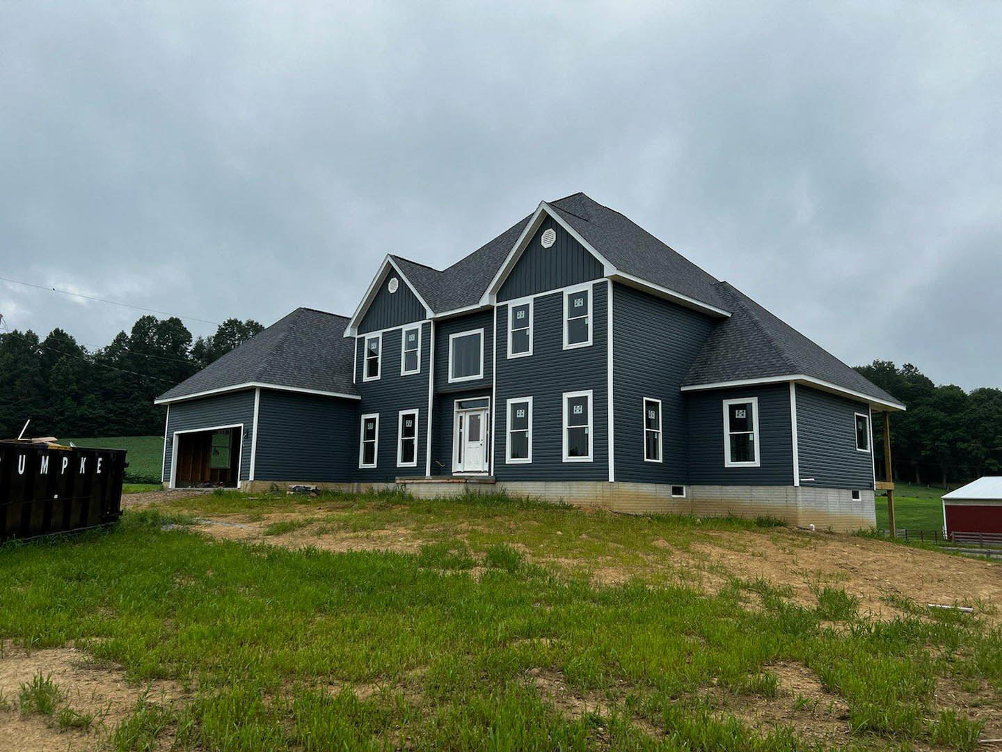 Two-story farmhouse with white siding and large windows, expansive green lawn with a white chair, black dumpster near driveway, overcast grey sky