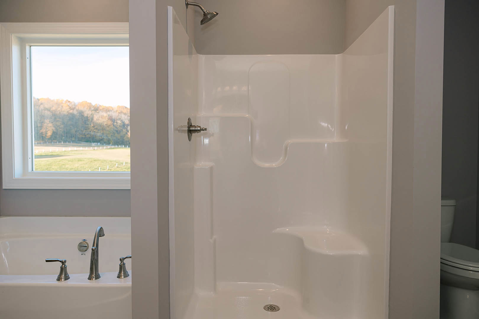 White tile shower with silver shower head and handle, window overlooking trees, adjacent toilet visible